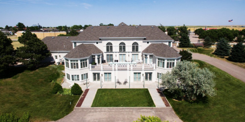 Aerial view of a large, elegant white mansion surrounded by green lawns and trees under a clear blue sky.