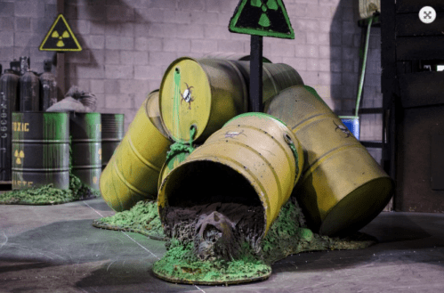 A pile of yellow barrels, some tipped over, with a figure partially emerging from one, set in an industrial environment.