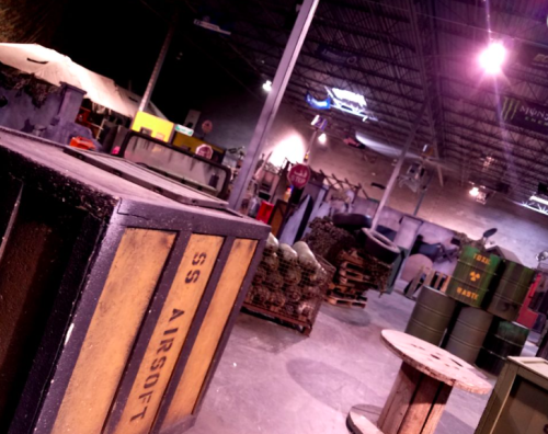 Indoor airsoft arena featuring various props, barrels, and crates in a dimly lit environment.