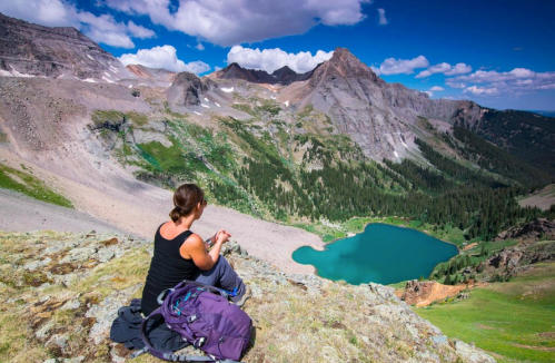 A hiker sits on a rocky ledge, overlooking a turquoise lake surrounded by mountains and lush greenery under a blue sky.