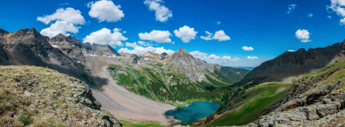 A panoramic view of mountains and a turquoise lake under a bright blue sky with fluffy white clouds.