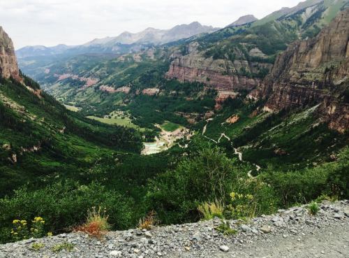 A scenic view of a lush green valley surrounded by mountains, with winding roads and rocky cliffs in the background.