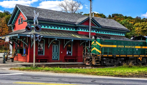 A green and yellow train passes by a red train station surrounded by autumn foliage under a blue sky.