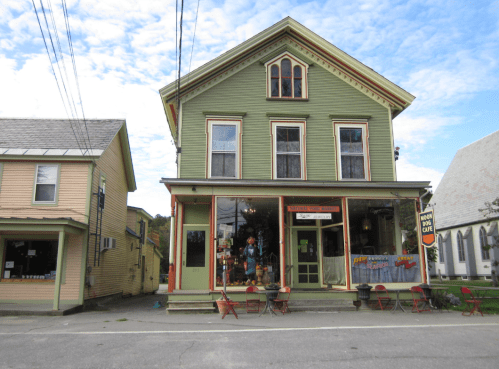 A green Victorian-style building with large windows, featuring a café and outdoor seating, under a cloudy sky.
