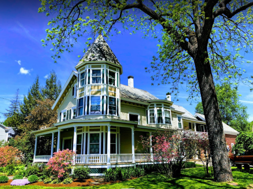 A charming two-story house with a turret, surrounded by lush greenery and colorful flowers under a clear blue sky.