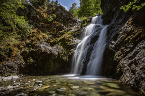 A serene waterfall cascades over rocky terrain into a clear pool, surrounded by lush greenery and sunlight.