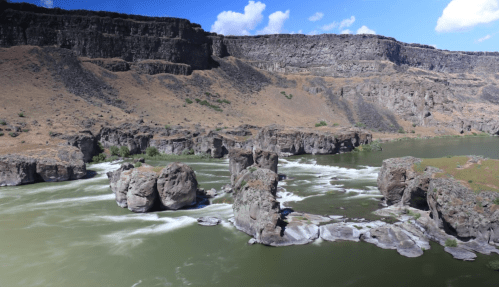 A scenic view of a river with rocky formations, surrounded by cliffs and dry land under a blue sky with clouds.
