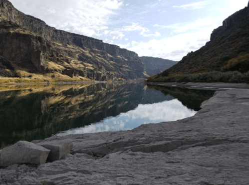 A serene river flows between steep cliffs, reflecting the sky and surrounding landscape on a calm day.
