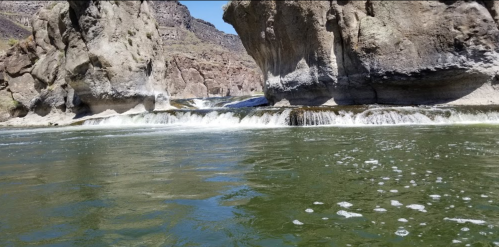 A serene river flows between rocky cliffs, with gentle waterfalls and bubbles on the water's surface under a clear sky.
