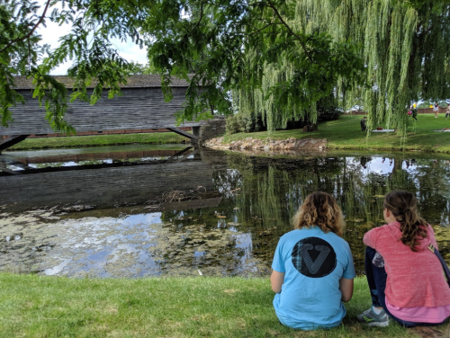 Two people sit by a pond, reflecting on the water, with a wooden structure and trees in the background.