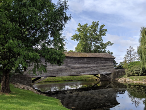 A wooden covered bridge spans a calm pond, surrounded by lush greenery and trees under a partly cloudy sky.