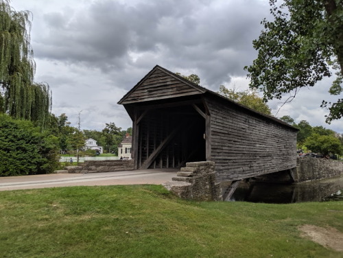 A wooden covered bridge spans a small stream, surrounded by greenery and under a cloudy sky.