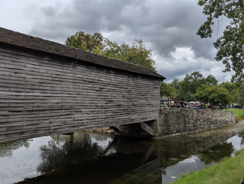 A wooden covered bridge spans a calm stream, surrounded by trees and a cloudy sky.