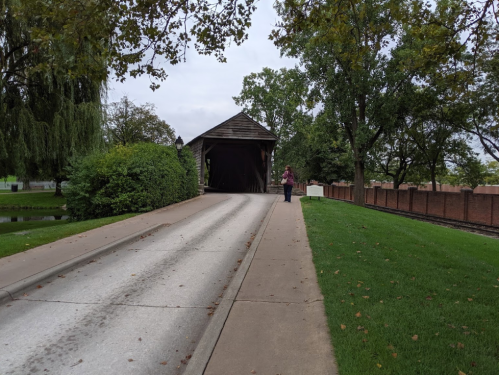 A person stands near the entrance of a covered bridge, surrounded by trees and a grassy area.