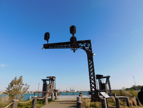 A metal structure with traffic lights stands over a pathway leading to a waterfront, under a clear blue sky.