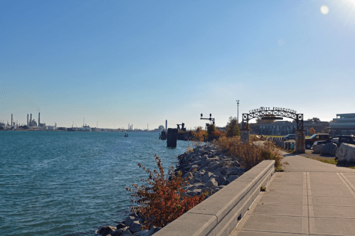 A scenic waterfront view with a pathway, industrial skyline, and colorful autumn foliage under a clear blue sky.