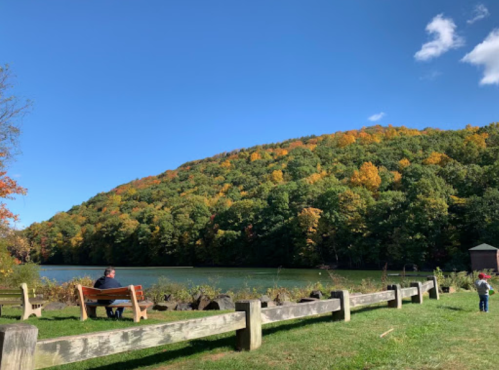 A serene lake scene with a man sitting on a bench, colorful autumn trees on the hillside, and a child playing nearby.