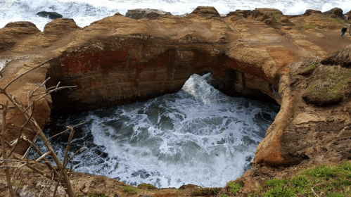 A rocky coastal arch with waves crashing below, surrounded by rugged terrain and greenery.