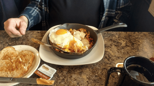 A skillet with a fried egg, vegetables, and meat, alongside toast and a cup of coffee on a table.