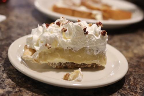A slice of pie topped with whipped cream and chopped nuts on a white plate, with a piece of bread in the background.