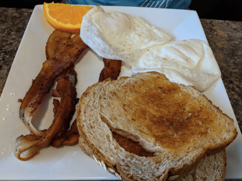 A plate with two poached eggs, crispy bacon, toasted bread, and a slice of orange.