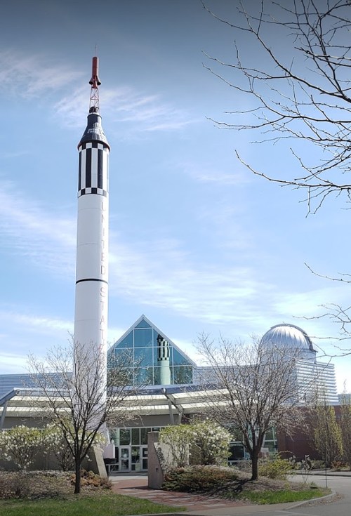 A rocket stands in front of a modern building with a dome, surrounded by trees and blue sky.