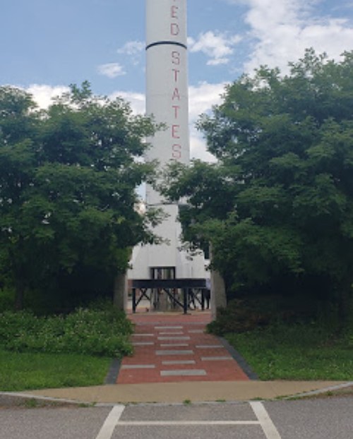 A tall rocket monument stands between two trees, with a pathway leading up to it under a partly cloudy sky.
