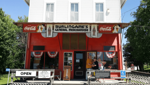 A vintage general store with a red facade, featuring Coca-Cola signage and two people standing at the entrance.