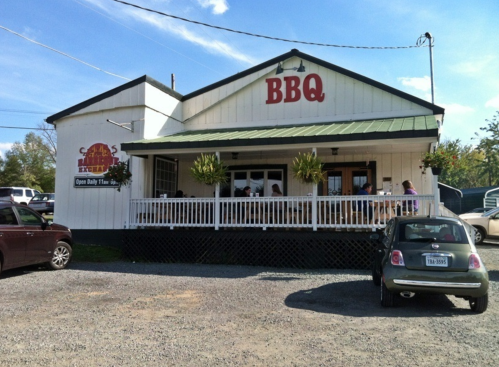 A BBQ restaurant with a white exterior, porch seating, and a sign indicating it's open daily. Cars are parked outside.