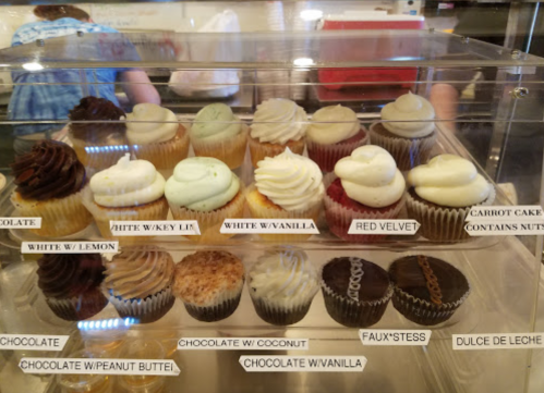 A display case filled with various cupcakes, labeled by flavor, including chocolate, red velvet, and coconut.