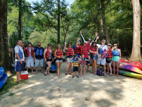 A group of people in life jackets poses together by kayaks in a wooded area, smiling and enjoying a sunny day.