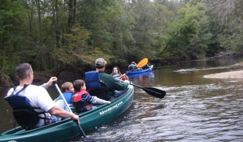 A family kayaking on a calm river surrounded by trees, with one kayak in the foreground and another in the background.