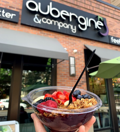 A hand holding a colorful acai bowl topped with granola and fresh berries, in front of a restaurant sign.