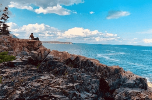 A person sits on rocky cliffs overlooking a serene blue ocean under a partly cloudy sky.