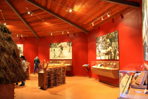 Interior of a museum exhibit with red walls, wooden beams, and displays of artifacts and historical images.