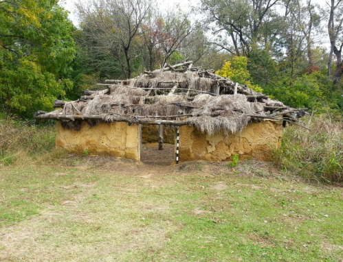 A rustic, thatched-roof structure made of mud and wood, surrounded by greenery and trees.
