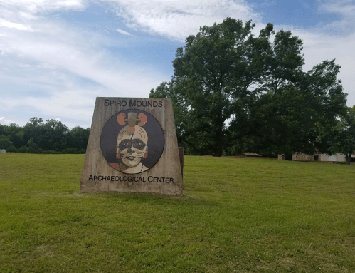 Sign for Spiro Mounds Archaeological Center, featuring a colorful depiction of a face, set in a grassy area.