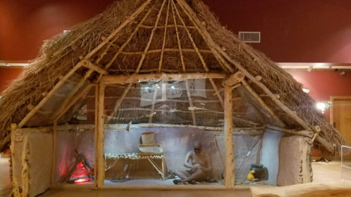 A traditional thatched hut interior with a figure sitting inside, showcasing historical living conditions.