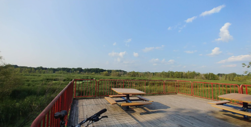 View from a wooden deck with picnic tables overlooking a lush green landscape and blue sky with scattered clouds.