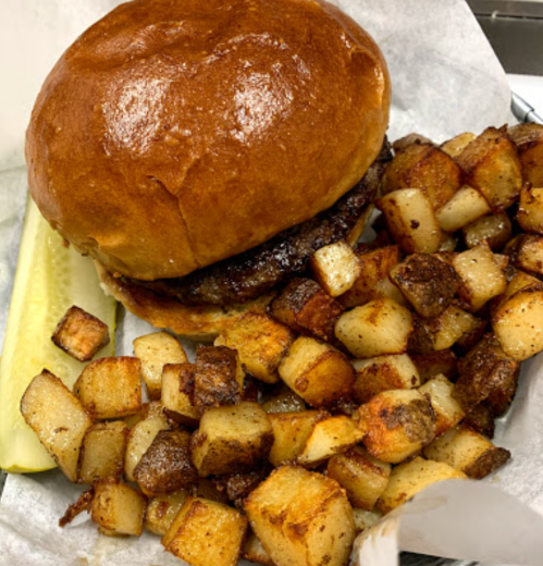 A juicy burger on a toasted bun with crispy diced potatoes and a pickle on the side, served in a paper-lined basket.