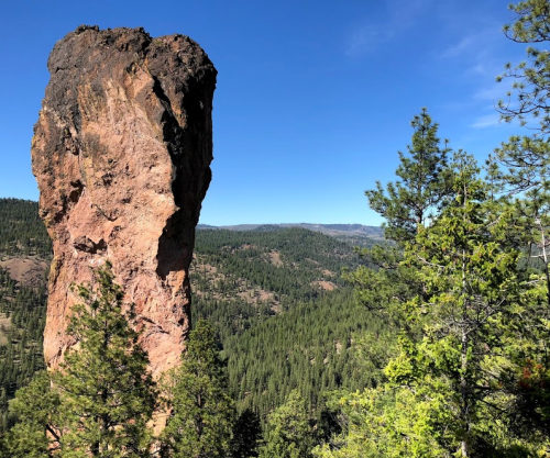 A tall rock formation stands prominently among green trees, with a clear blue sky and distant mountains in the background.