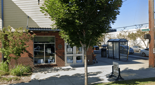 A brick building with large windows, a bus stop nearby, and trees lining the sidewalk on a sunny day.