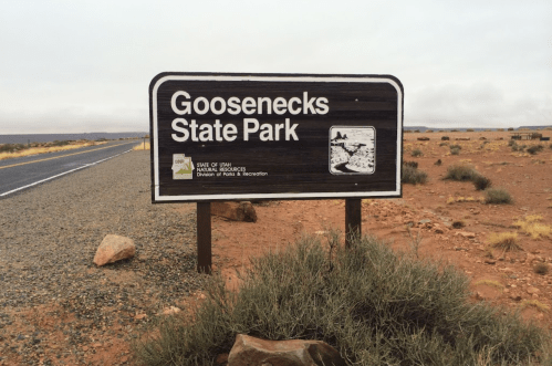 Sign for Goosenecks State Park along a road, with a desert landscape and cloudy sky in the background.