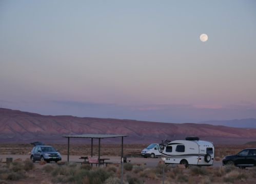 A serene desert landscape at dusk with a full moon, parked cars, and a picnic area under a shelter.