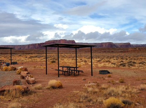 A picnic area with a table and shelter in a desert landscape, surrounded by dry grass and distant rock formations under cloudy skies.