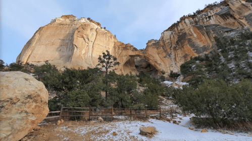 A rocky cliff with a large arch, surrounded by trees and a snow-dusted ground, under a clear blue sky.