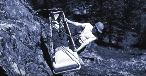 A climber maneuvers a stretcher along a rocky cliffside, secured with ropes and wearing a helmet.