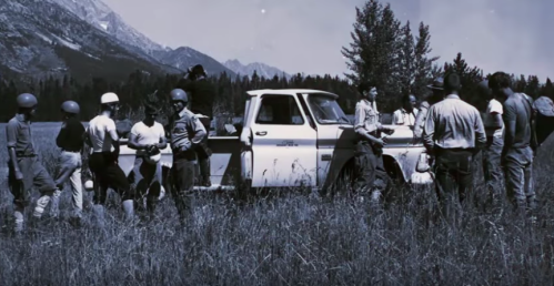 A group of people in helmets gathered around a white truck in a grassy field with mountains in the background.