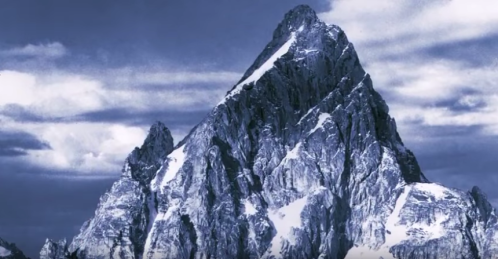 A majestic mountain peak with snow-capped edges under a cloudy sky, showcasing rugged terrain and dramatic shadows.