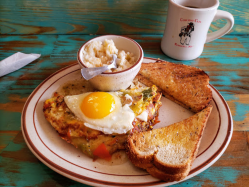 A plate with a vegetable omelet topped with a fried egg, a bowl of grits, and two slices of toasted bread, with coffee.